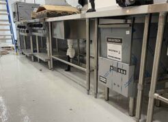 A commercial kitchen under installation, featuring stainless steel sinks, countertops, and a Washtech dishwasher, with a ladder and electrical cords in the background.
