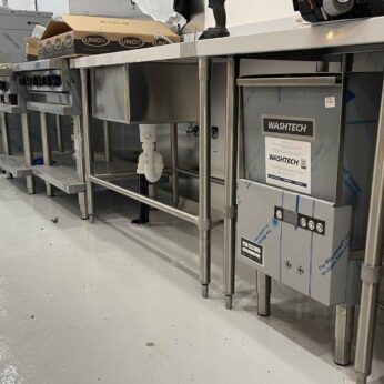 A commercial kitchen under installation, featuring stainless steel sinks, countertops, and a Washtech dishwasher, with a ladder and electrical cords in the background.