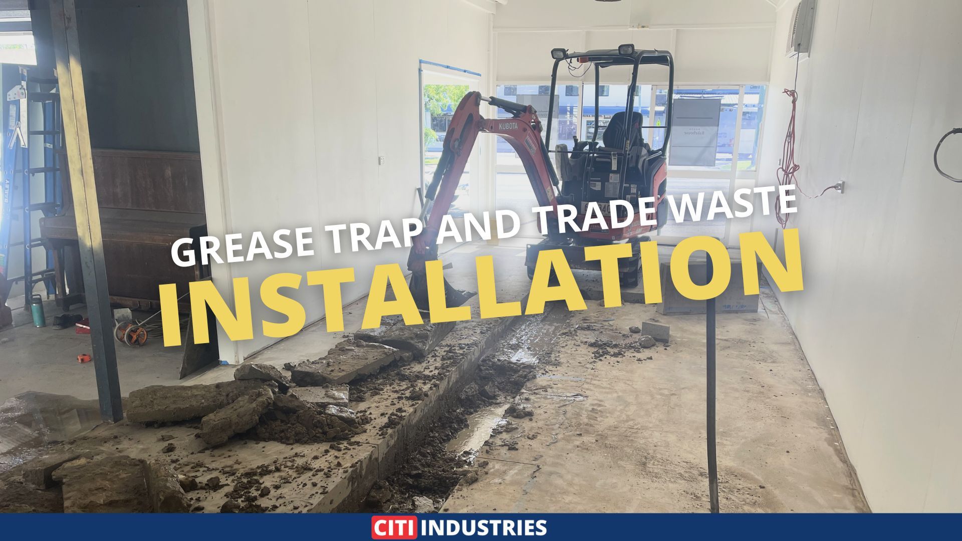 An excavator digs a trench through a concrete slab inside a Sarina commercial tenancy, with broken concrete and exposed earth visible along the cut line.